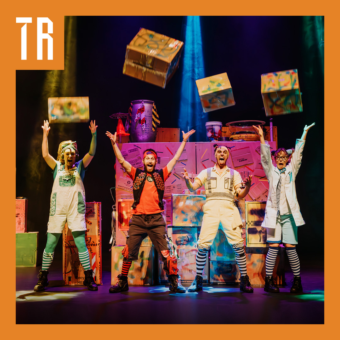 Four performers stand on a stage, they are brightly dressed, and look raucous. They each throw a cardboard box into the air