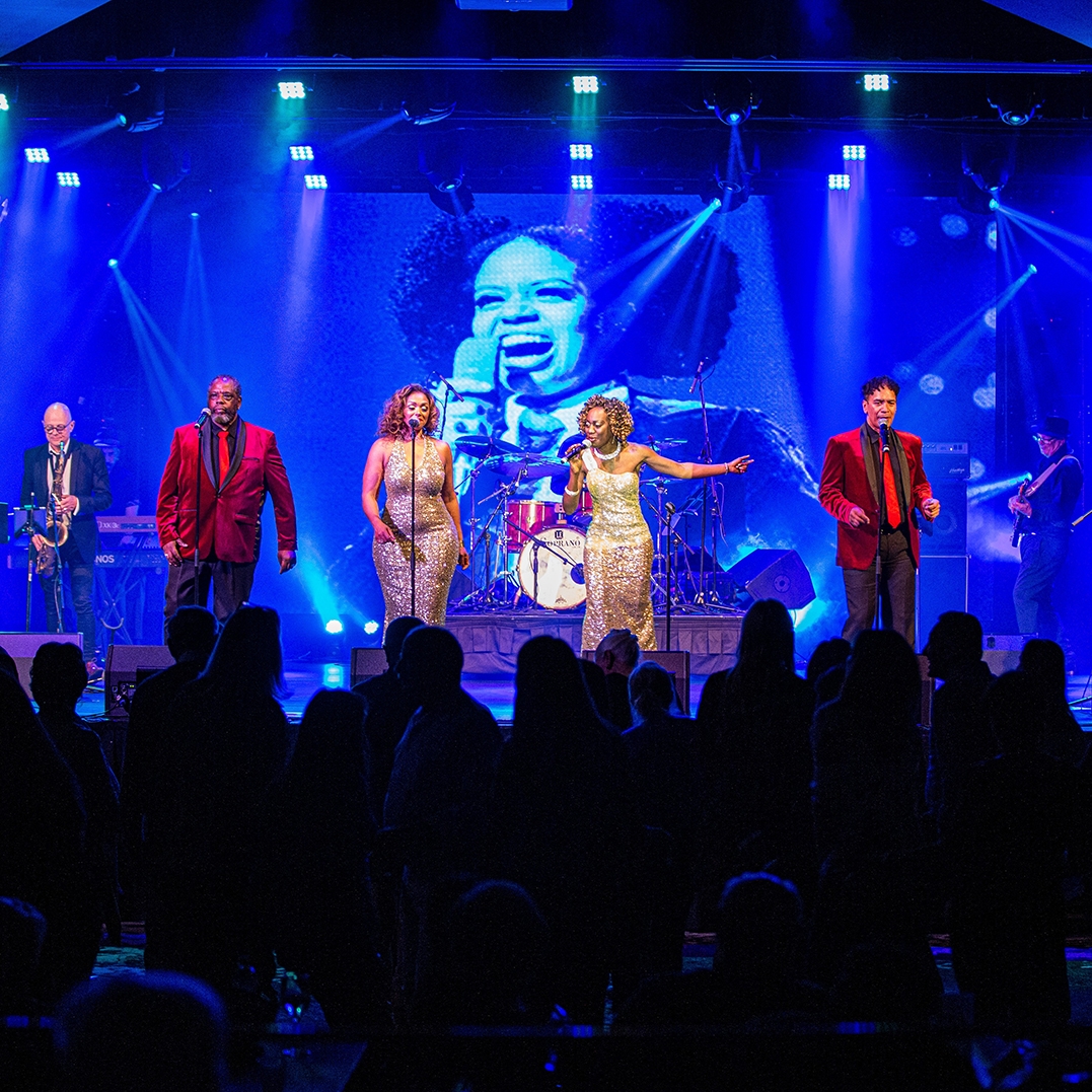 Four glamourous singers perform on a ble lit stage, in the background there is a projection of Diana Ross. The female singers wear sparkeling dressed the two male singers wear red satin jackets