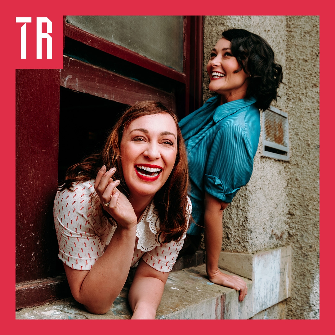 Two woman in 1950 style clothing and hairstyles lean out of a window smiling