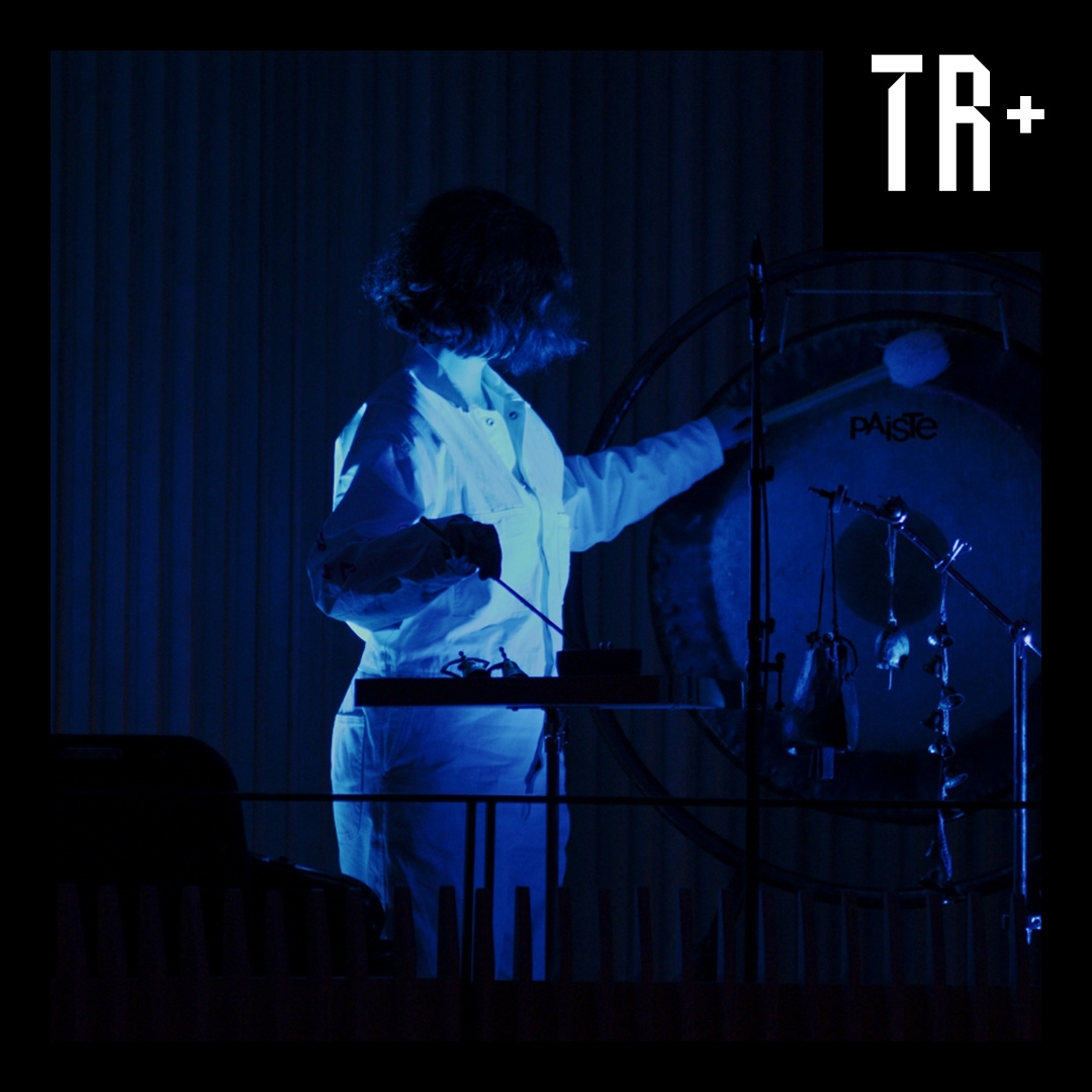 A dark immage illuminated in blue of a musician weaing while playing percussive instruments