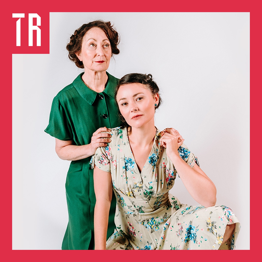 Two women in 1950s clothing and style. A younger woman sits in the front in a floral dress with a motherly figure behind her gently resting her hands on her sholders.