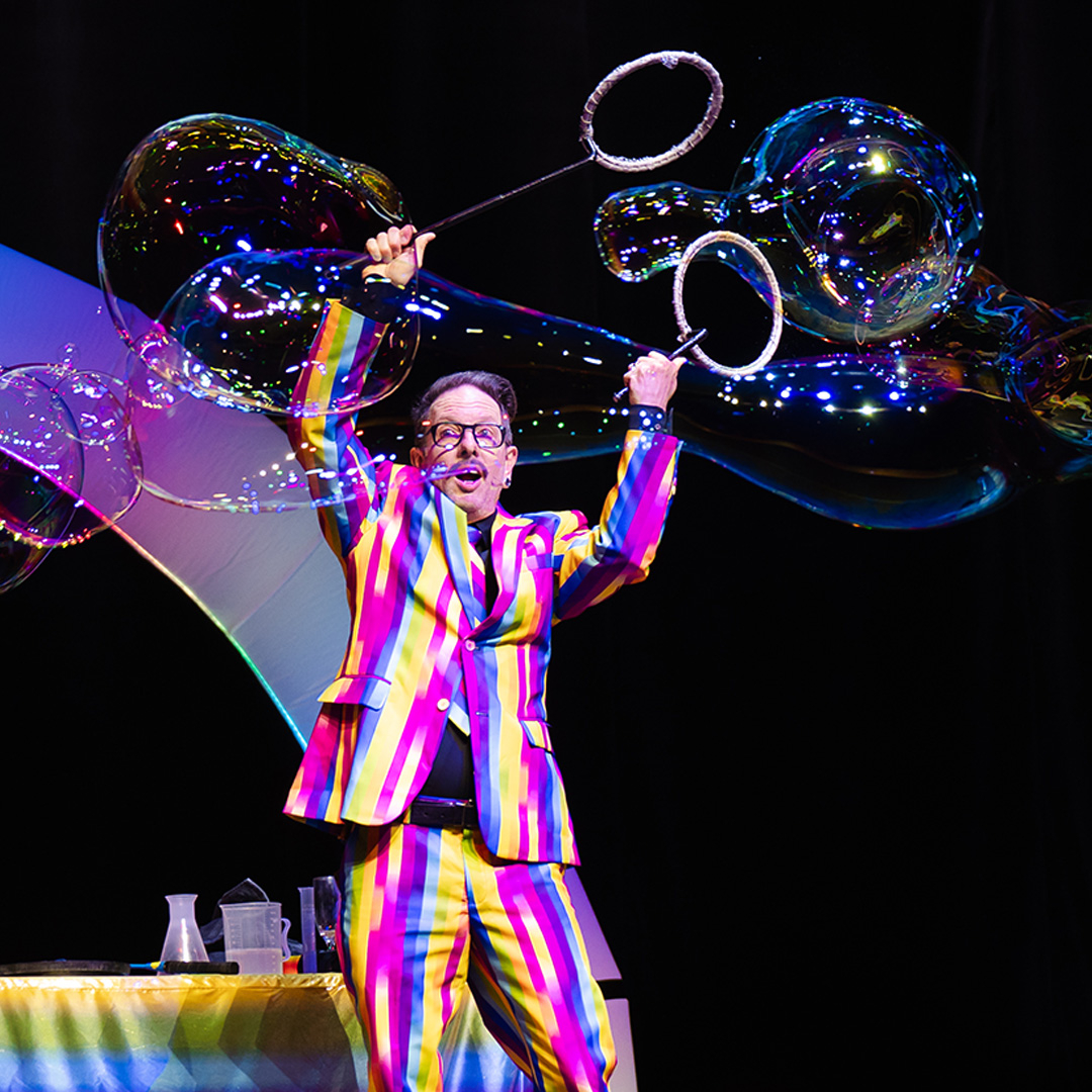 A man wearing a colourful stripey suit does tricks with giant bubbles