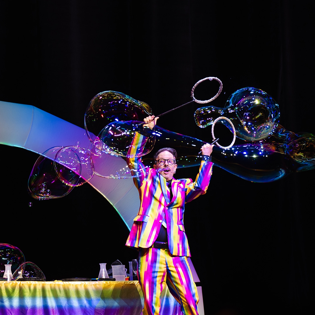 A man wearing a colourful stripey suit does tricks with giant bubbles