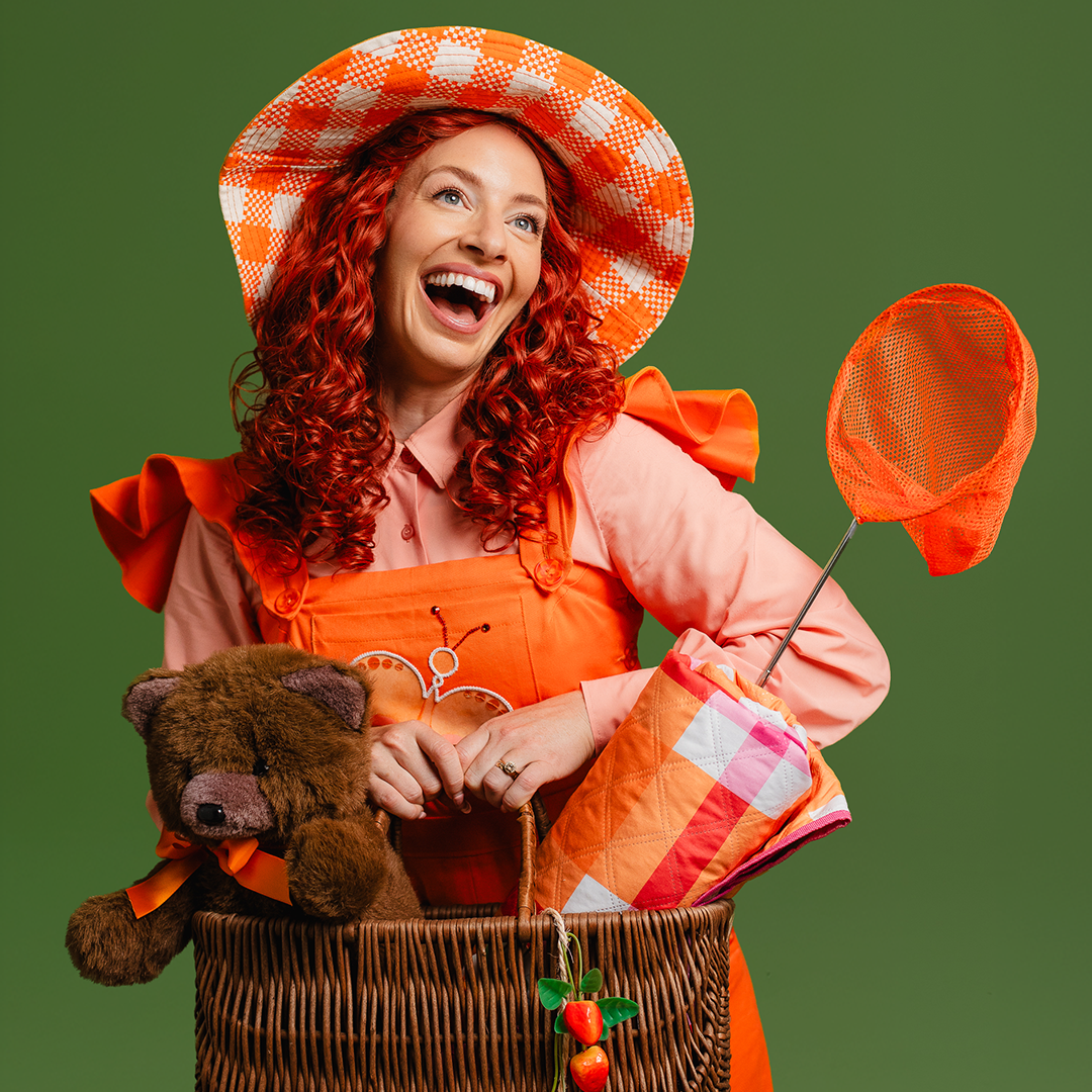 A portrait of childrens performer Emma Memma, she is wearing bright orange clothing and holds a basket with a teddy, fishing net and picnic mat