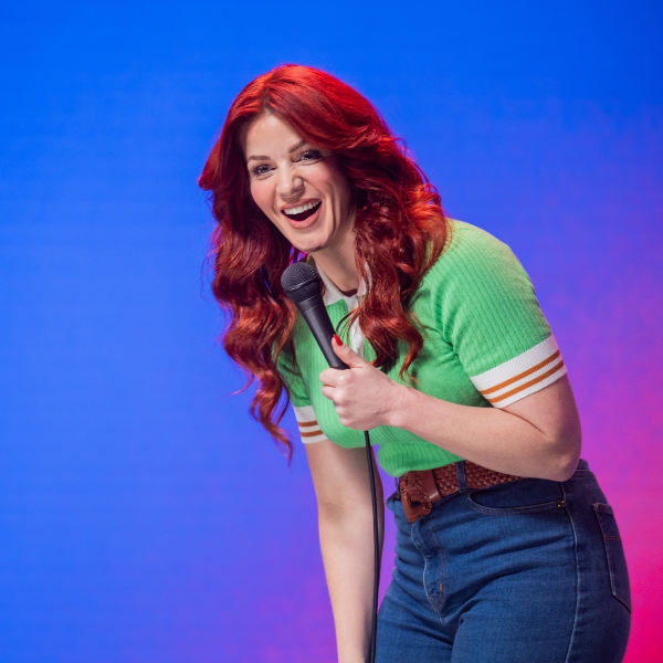 A portrait of comedian Bron Lewis. She is wearing bright retro clothes and has bright red hair