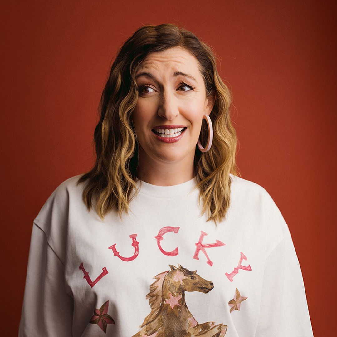 Portrait of comedian Celia Pacquola wearing an 80s wincheater with a horse and the word lucky in red.