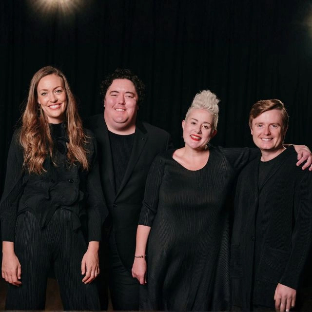 A portrait of singers Katie Noonan, Hannah Fraser, Louis Hurley, and Andrew O’Connor. They all wear black and smile. It impression is stylish but welcoming