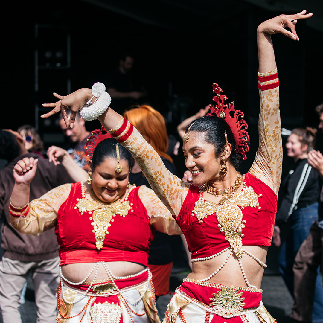 Two female Indian Traditional dancers wearing traditional dress in red and gold pose smiling, mid dance