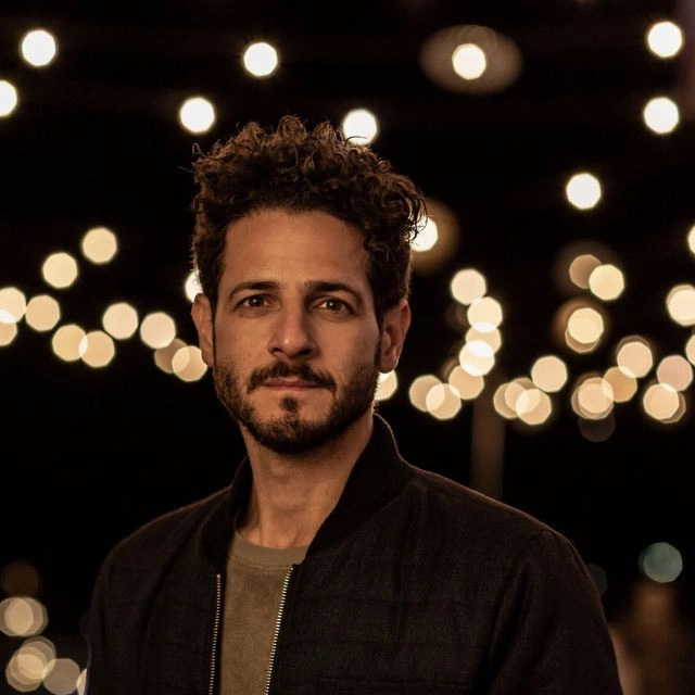 A portrait of singer-songwriter Lior. A mand with curly short hear and a bears stands againsta. drak background illuminated by gold lights