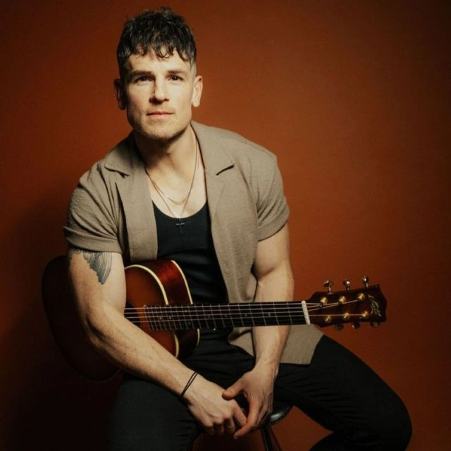 A portrait of singer Michael Paynter, he sits on a stool leaning slightly forward, with his guitar tucked under his arm. The colours are red, tan and earthy