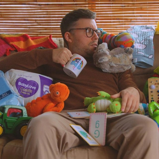 A portrait of Rumple, a dad man is sitting on a couch holding a coffee mug covered in toys and toddler bric a brac