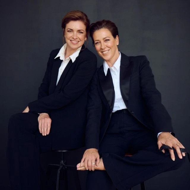 A portrait of singers Gorgi Coghlan and Anna-Lee Robertson, both dressed in black suits, with a white shirt, no tie
