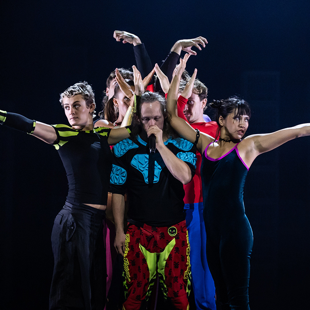 A group of dancers in bright sporty fun costumes pos one stage. One is holding a microphone.