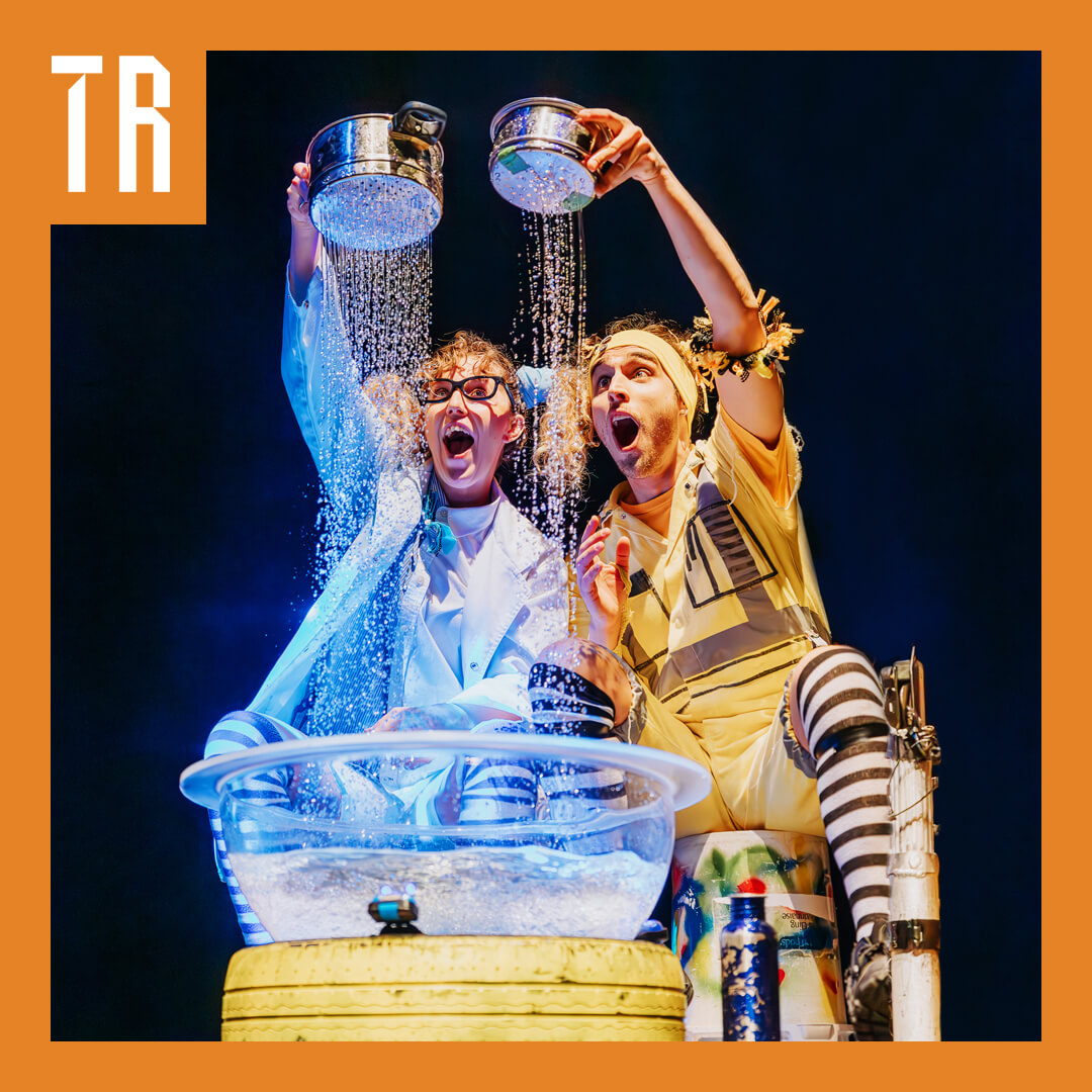 Two people hold pots in the air which have water in them. The pot have holes and there is water from the pots raining down. The two performers look on amazed by this.
