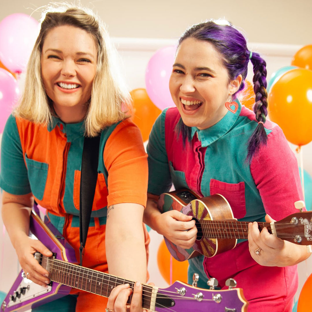 A colourful protrait of two female musicians one holding a guitar, one a ukelele. They are dressed in colourful clothes with colourful balloons surrounding them