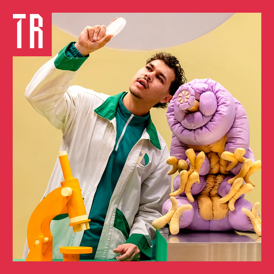 An actor dressed as a scientist is holding a pietrie dish up to the light, next to him a puppet tardigade, the size of a dog, looks on