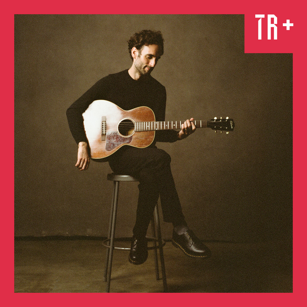 Portrait of guitarist Julian Lage, A man wearing black sits on a stool holing an accostic guitar, The image is earthy colours