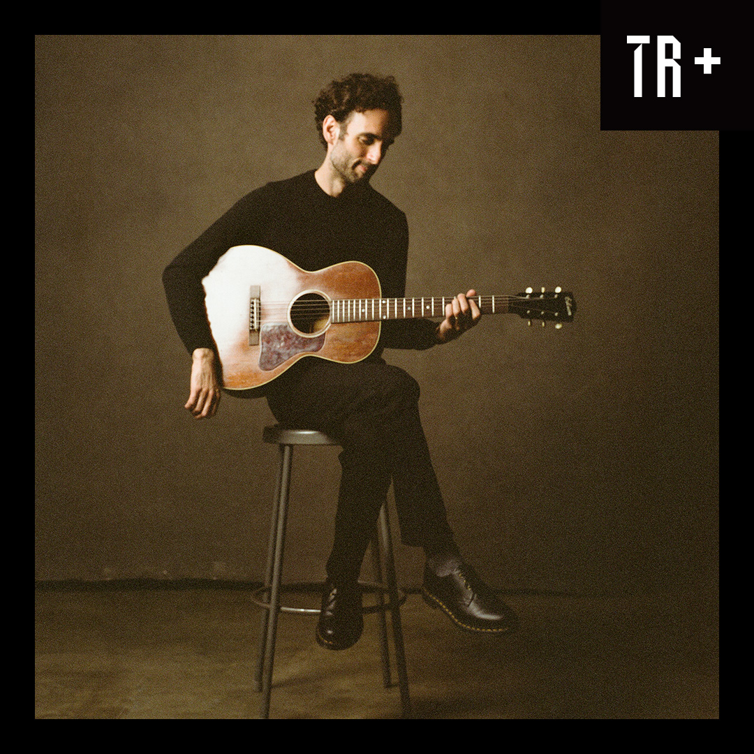Portrait of guitarist Julian Lage, A man wearing black sits on a stool holing an accostic guitar, The image is earthy colours