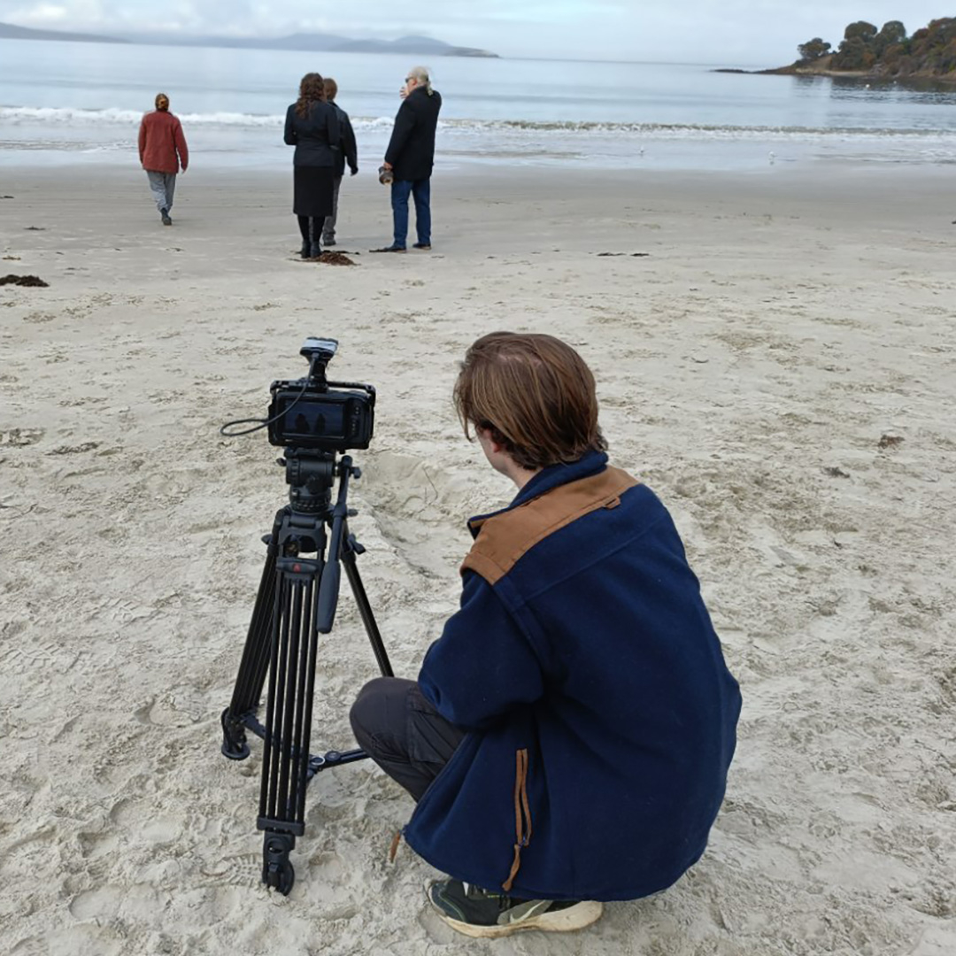 a film maker squats on a beach behind a camera set up on a tripod, there are people in the background