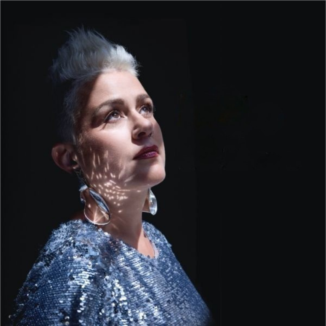 A close up portrait, Katie stands in the shadows with a bring sun light illuminating her face and sholders. She has platinum white hair and is waerinc  colourful clothes and earings in blue and silver tones