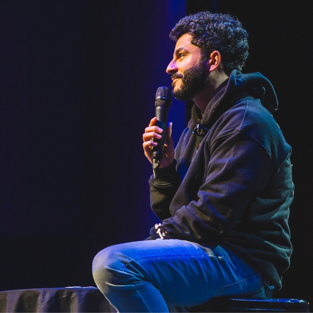 Photos of comedian Khaled Khalafalla , wearing a dark hoody and blue jeans, holding a mic sitting on a stage. He has short curly dark hair and a dark beard