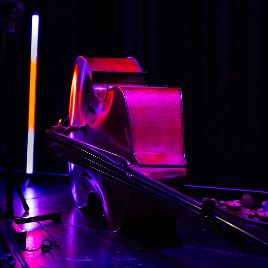 A cello illuminated by vivid purple and red stage lighting, resting on a dark stage beside a vertical light bar.