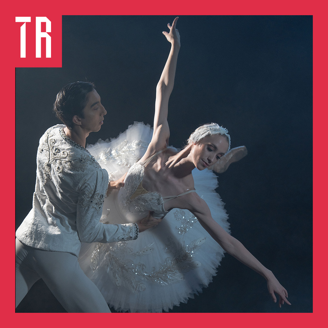 Two ballet dancers in white costumes perform a dramatic pose from Swan Lake on a dark stage, with the ballerina extending one arm upward and the other outward. A red border and white “TR” lettering frame the image.