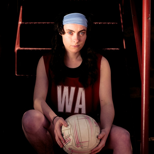 A young woman looks towards the camera wearing a netball bib and holding a netball.