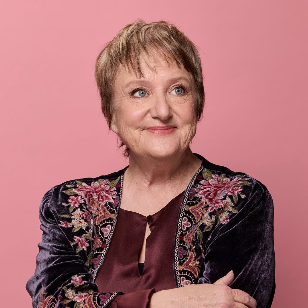 Portrait of Australian comedian Denise Scott , short light-brown hair, smiling gently at the camera. She wears a burgundy top and a dark velvet jacket with colorful floral embroidery, against a pink background.