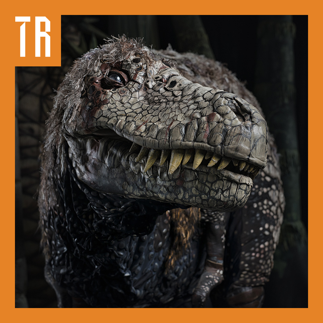 Close-up of a detailed dinosaur-like creature with scaly skin, sharp teeth, and a feathered head, facing the camera against a dark forest background.
