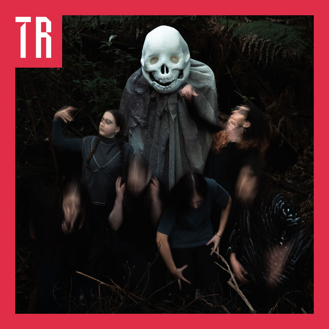 A dark forest scene featuring a large figure in a white skull mask surrounded by five blurred, dark-clothed figures, framed by a bold red border with white “TR” lettering in the upper-left corner.