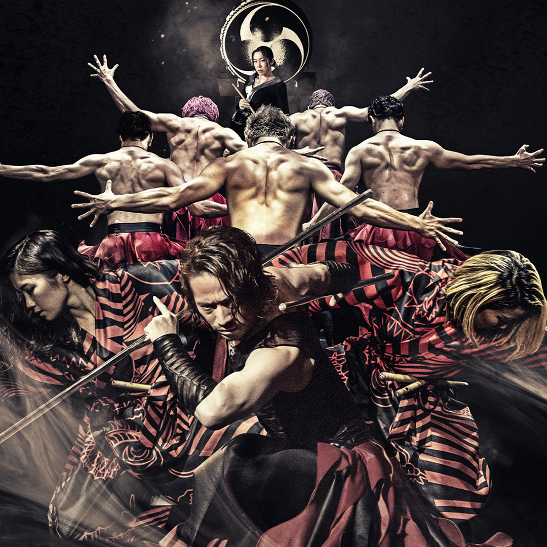 A dramatic stage performance featuring a group of dancers in black-and-red costumes, with shirtless dancers forming a raised formation behind them and a woman standing beneath a large circular emblem in the background.