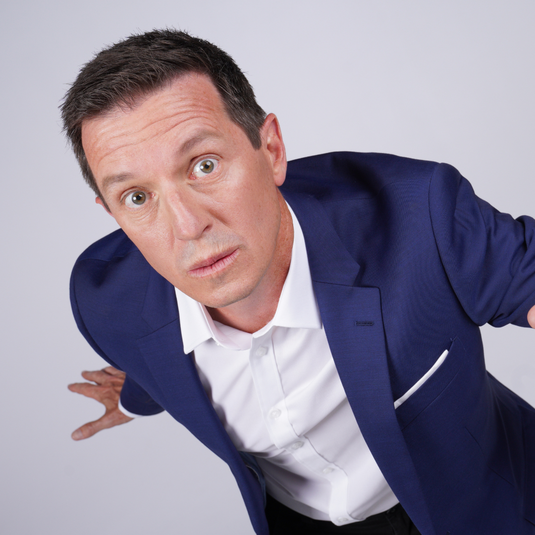 A man in a blue suit and white shirt leans toward the camera with a surprised expression.