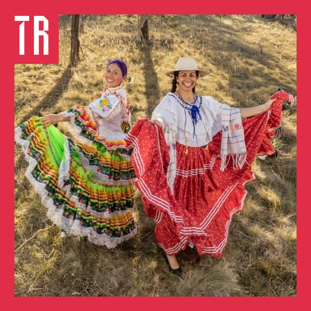Two smiling women pose outdoors in traditional folkloric dresses, holding out their colorful, ruffled skirts. One wears a white embroidered blouse with a green, yellow, and red skirt, while the other wears a white blouse, straw hat, and red skirt with white trim. They stand on a sunlit, grassy hillside, framed by a bold red border with a white “TR” logo in the upper-left corner.