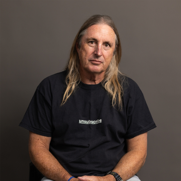 A middle-aged man with long, straight, light brown hair sits facing the camera against a plain grey background. He wears a black T-shirt with “unsubscribe” printed on the chest, light-coloured trousers, and a wristwatch. His hands rest in his lap, and he has a neutral expression.