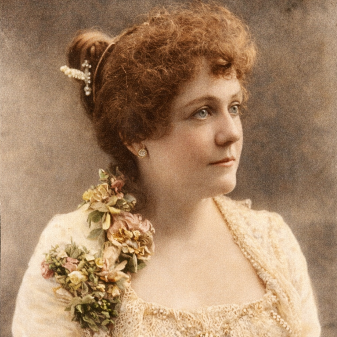 A vintage, colorized portrait of a young woman with auburn hair styled in an elaborate updo adorned with pearl ornaments. She wears pearl earrings and a cream lace dress with a floral arrangement of pink, yellow, and white flowers draped over her shoulder, gazing thoughtfully to the right against a softly textured studio backdrop.