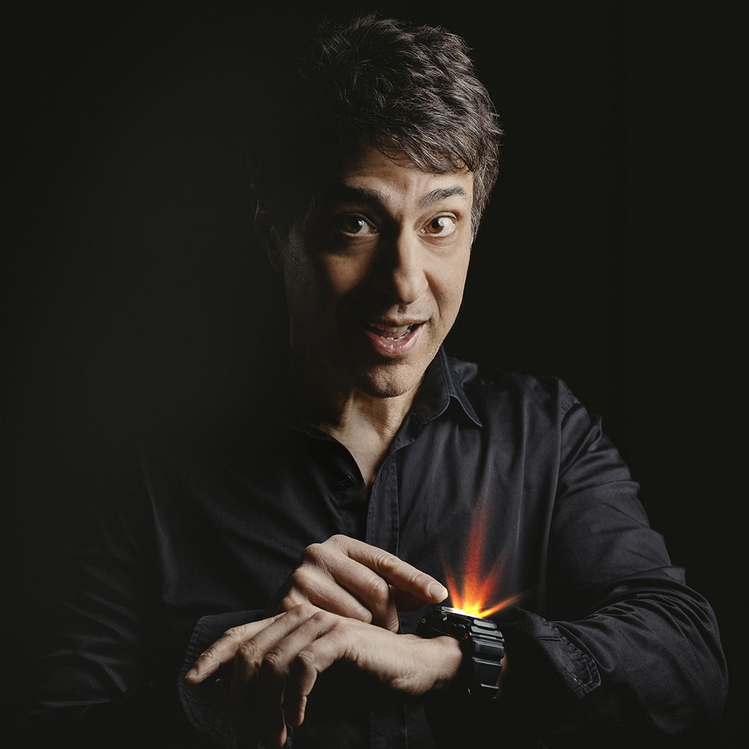 Portrait of comedian Arj Barker - A man in a black shirt looks surprised as he points to his wristwatch, which glows with a bright, fiery orange light. Dramatic low-key lighting illuminates his face and hands against a dark background, creating a mysterious, cinematic atmosphere.