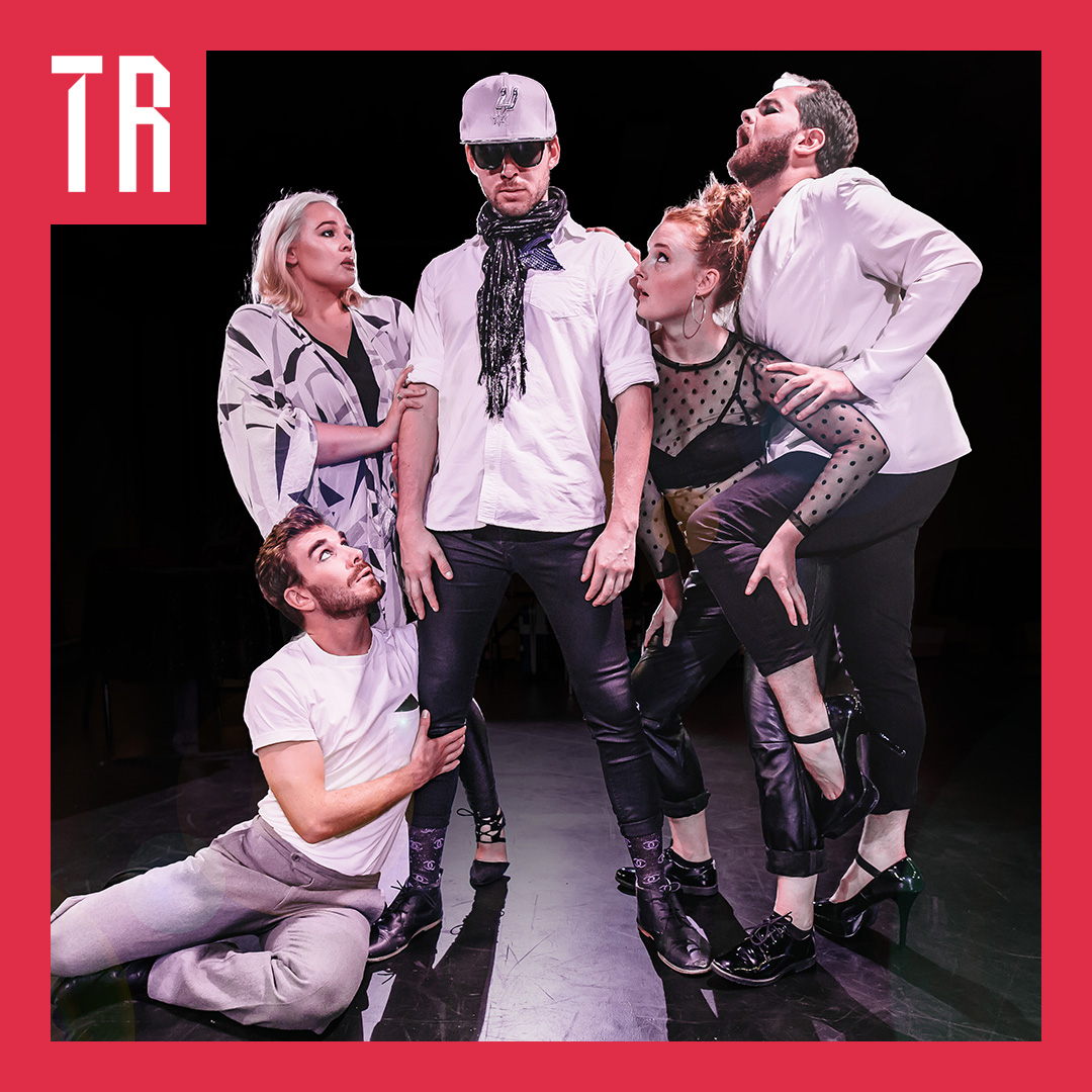 A group of five performers pose dramatically on stage in white tops, dark trousers and high heels, with one man wearing a cap and sunglasses at the centre. Two performers crouch at his feet while the others lean towards him, creating a theatrical, humorous tableau. The photograph is framed by a bright red border with a white “TR” logo in the top-left corner.
