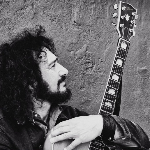 A black and white image of a person holding a guitar. His head is turned to his left. He stands in front of a rock face.