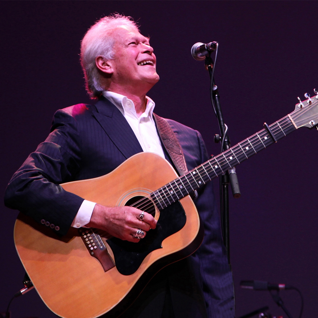 Photo: supplied by the artist A man with grey hair looks towards the sky. He stands in front of a microphone on a stand and holds a guitar.