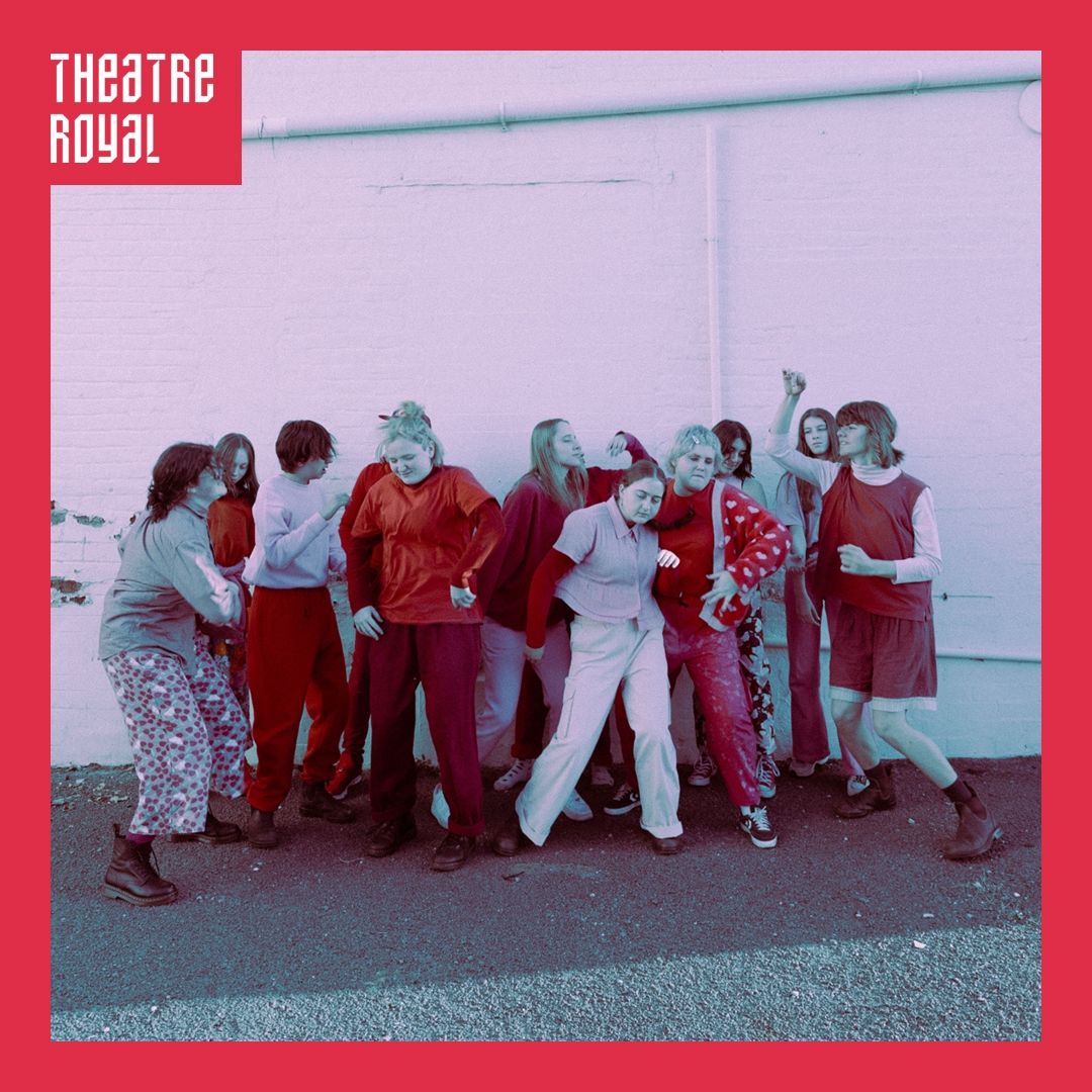 Photo: Darcie Richards A group of young dancers stand close together. They are outside against a white, industrial looking wall. They are wearing shades of pink and red.