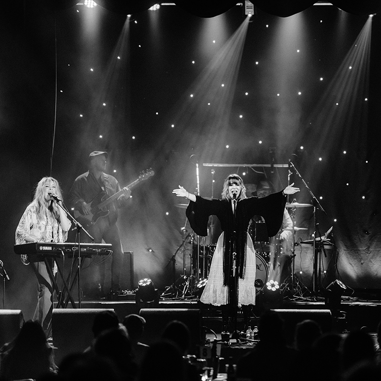 A black and white image of a full band on stage. The singer has their arms outstretched and there are lights shining down on them.
