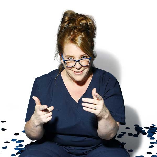 A woman sits on the floor in a nurses blue uniform. Her hair is up and she wears glasses. She is surrounded by blue confetti.