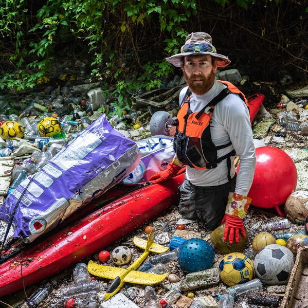 A man in a hat and life jacket on his knees next to a red kayak surrounded by rubbish.