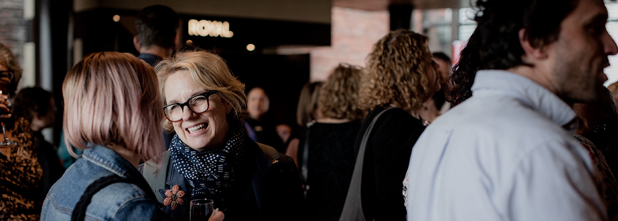 A crowd gathered in the foyer of the Theatre Royal. A woman with glasses smiles in the foreground.