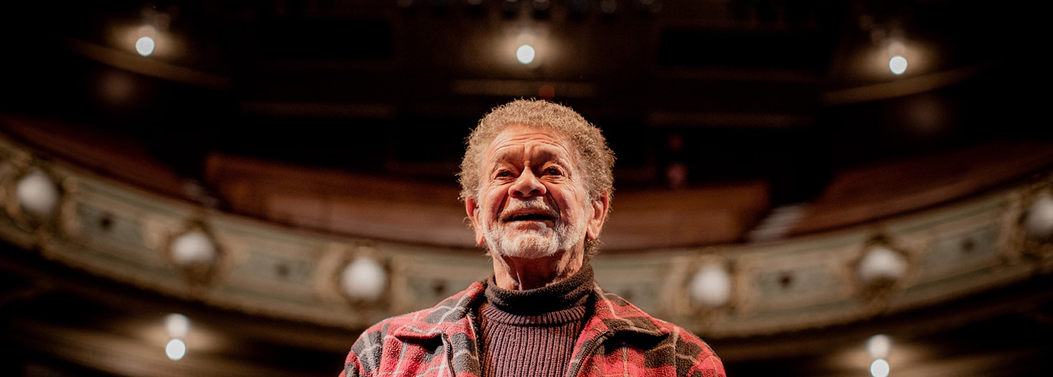 Image: Rosie Hastie An Aboriginal man, Uncle Jim Everett – puralia meenamatta stands on the Theatre Royal stage. He is smiling. Behind him is a seating of the theatre.