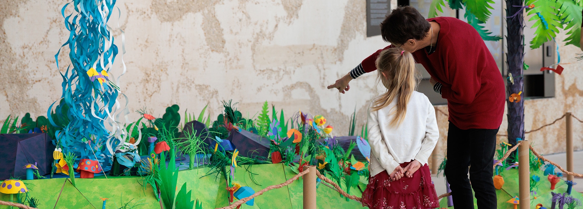 A child and an adult stand in front of a colorful environment made out of papaer. The adult is pointing to something in the scene